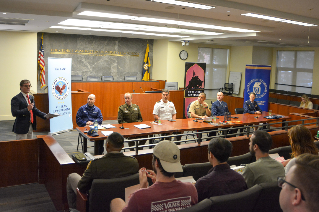 military personnel sitting at a table looking at the moderator speaking with a microphone to a crowd of students