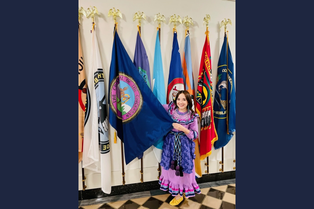 Morgan Gray, pictured in traditional Chickasaw regalia in front of the Chickasaw Nation flag