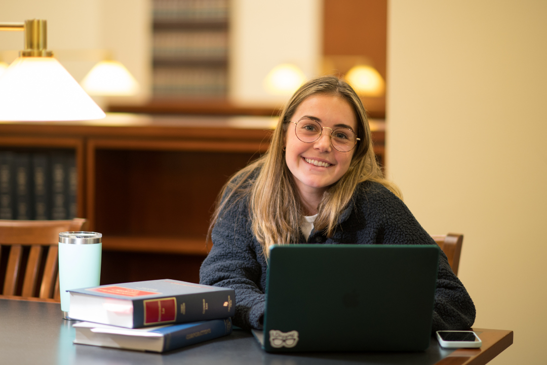 A female student sitting in the library with a laptop and books smiling