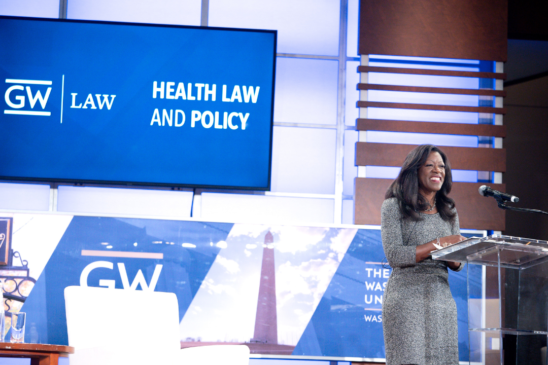 Dean Dayna Bowen Matthew behind a podium smiling with a digital screen in the back reading GW Law Health Law and Policy