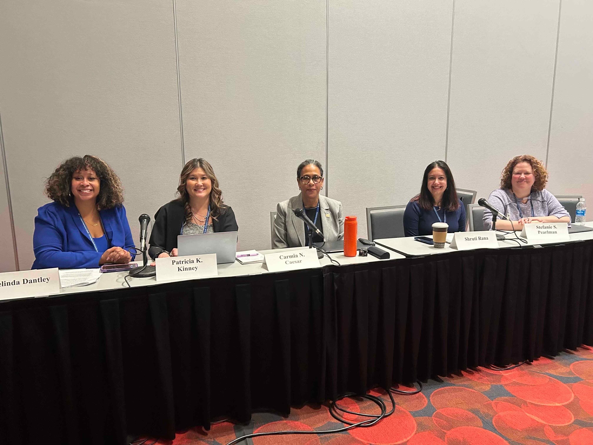 GW Law Associate Dean Carmia Caesar sits behind a black table during a panel at the 2025 AALS meeting in San Francisco. She is surrounded by a group of other law professors. 