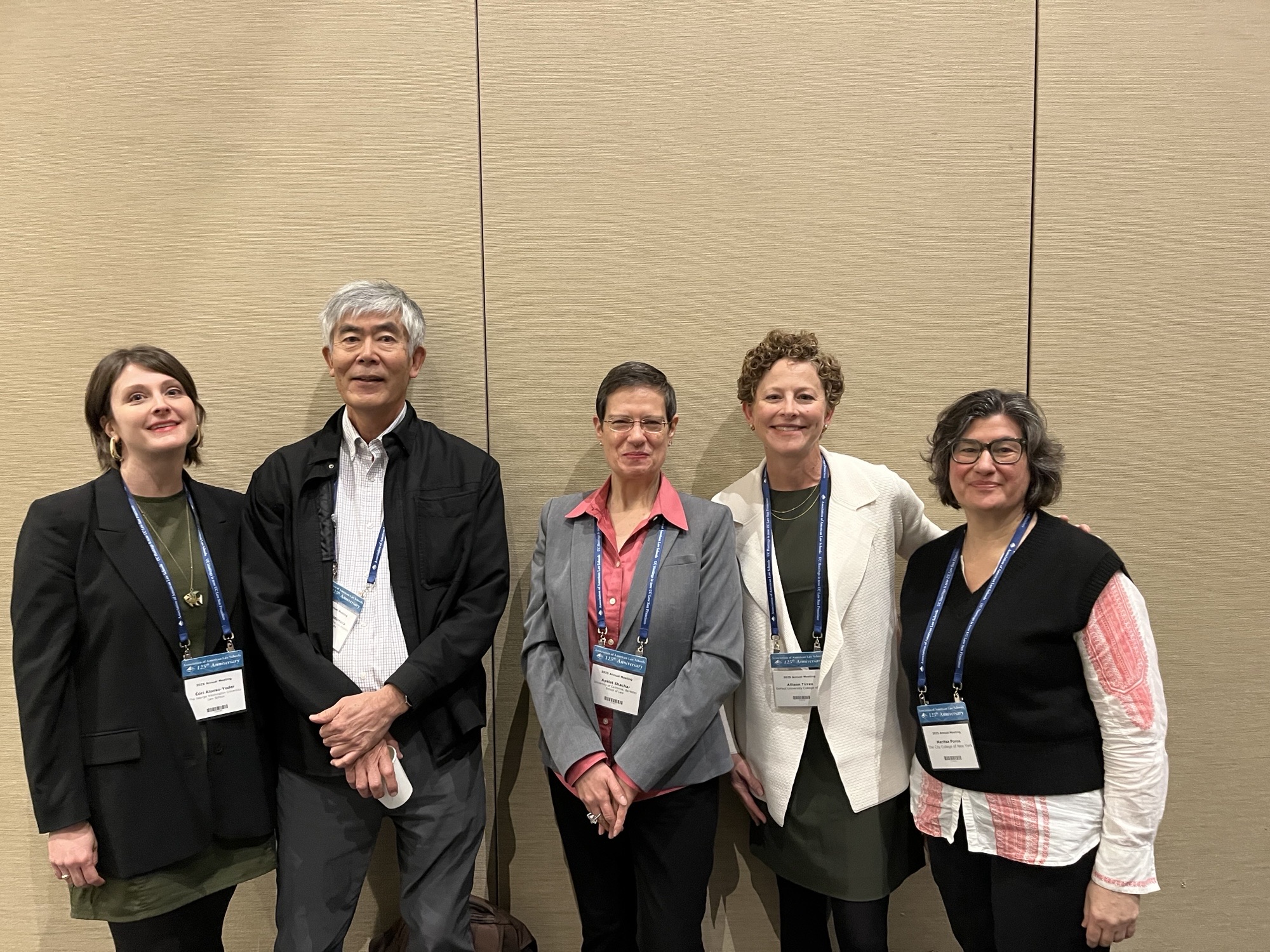 Cori Alonso-Yoder, Hiroshi Motomura, Ayelet Shachar, Allison Tirres, and Maritsa Poros pose for a photo in front of a beige wall during the 2025 annual AALS meeting.