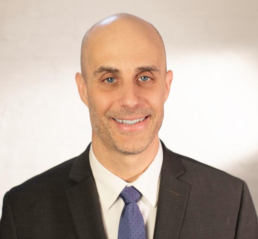 Headshot of Brian Tronic. He is wearing a black suit, white shirt, blue tie, and is smiling.