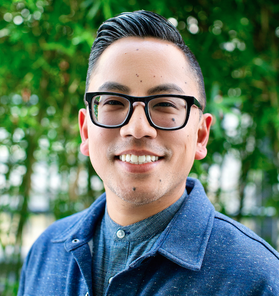 Headshot of Angelo Mathay. He is outside with trees i the background. He is wearing a blue button down, black-framed glasses, and is smiling.