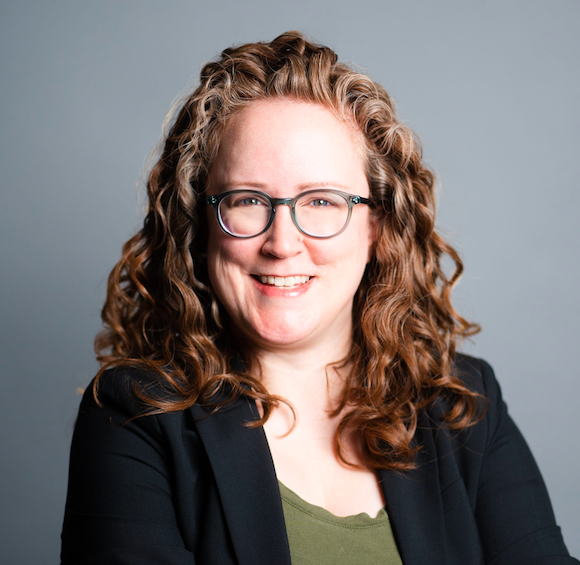 Headshot of Bridget Geraghty. She is wearing a green top, black blazer, and glasses. Her reddish-brown curly hair is down. Her arms are crossed and she is smiling..