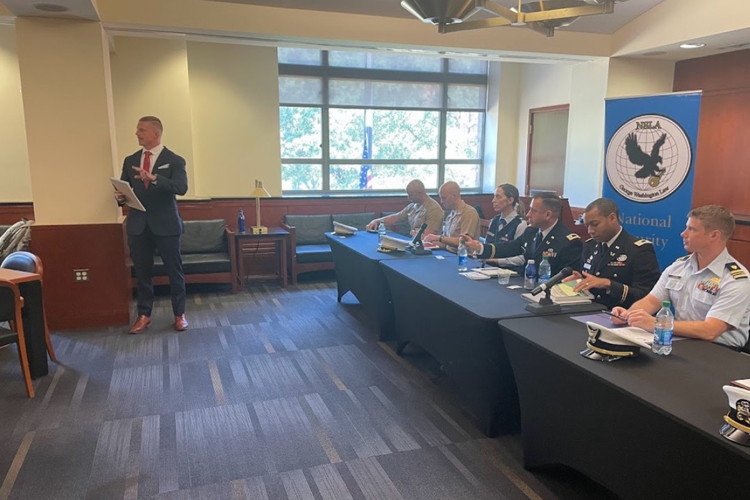 A group of military people sitting on a panel and a man standing up addressing the crowd