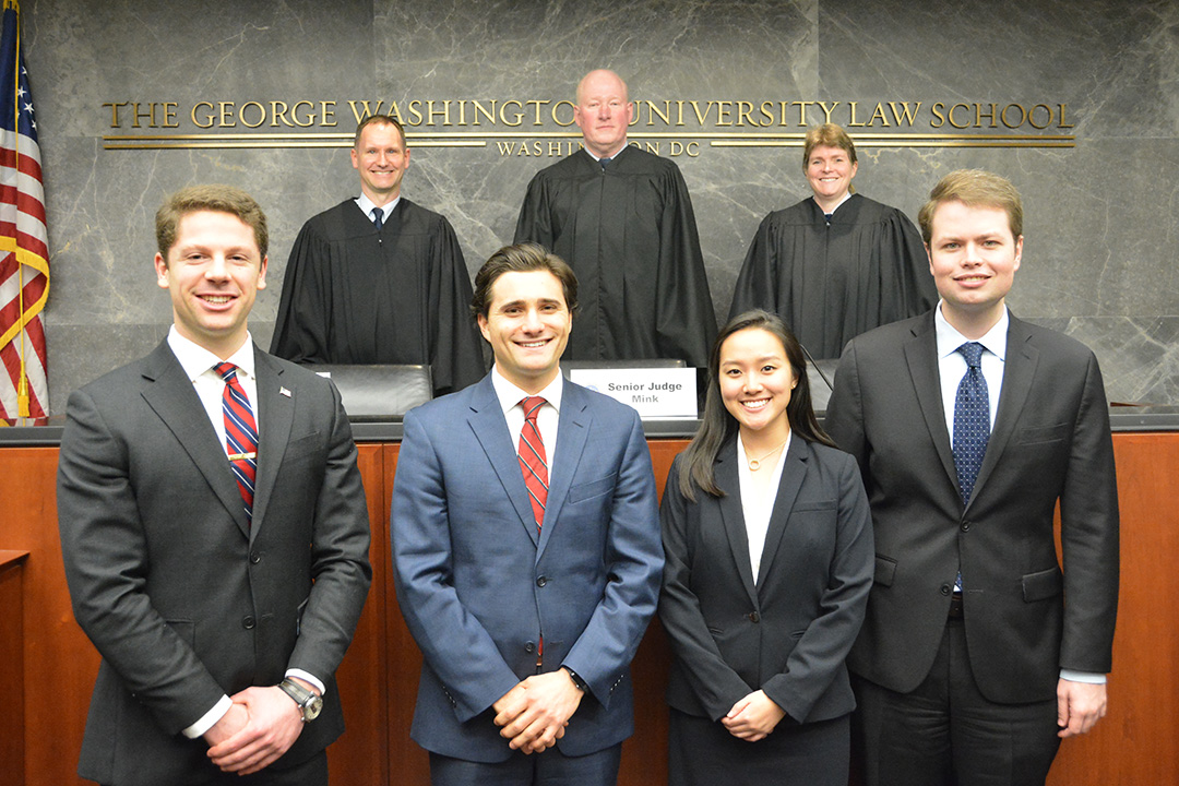 Three JAG judges stand behind the bench, with four GW Law students standing in front for a group photo.