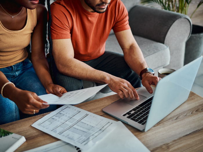 A black woman holding a piece of paper and a black man sitting on the couch looking at a laptop together