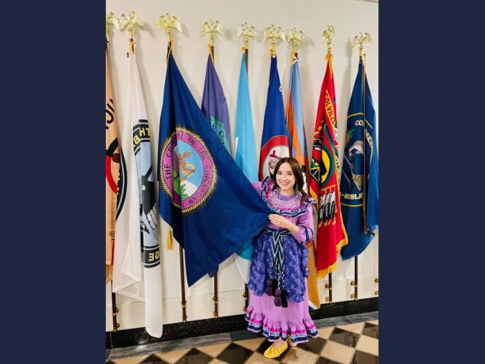 Morgan Gray, pictured in traditional Chickasaw regalia in front of the Chickasaw Nation flag