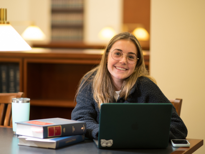 A female student sitting in the library with a laptop and books smiling