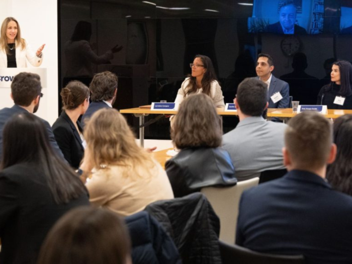 Jessica Tillipman speaking at a podium with a panel of 3 lawyers sitting to the right and a crowd in the foreground