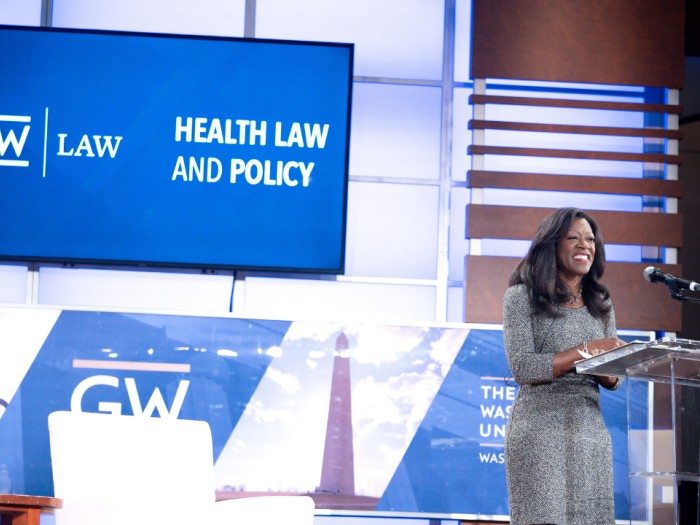 Dean Dayna Bowen Matthew behind a podium smiling with a digital screen in the back reading GW Law Health Law and Policy