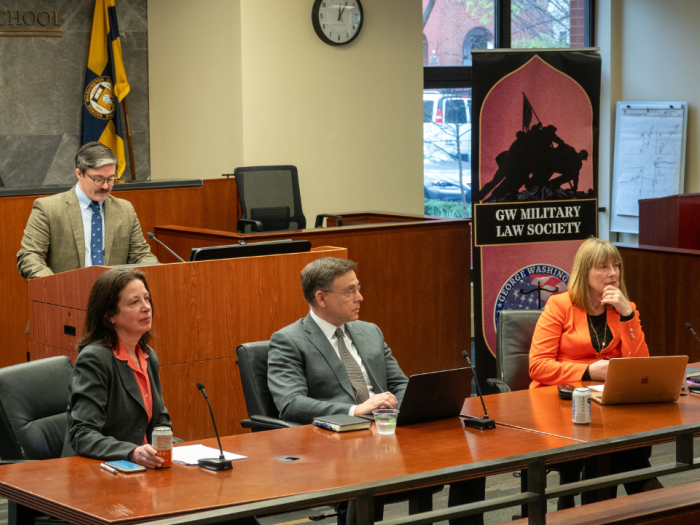 Professor Dickinson, Corn, and Ni Aolain sitting on a panel with a person behind the lectern talking