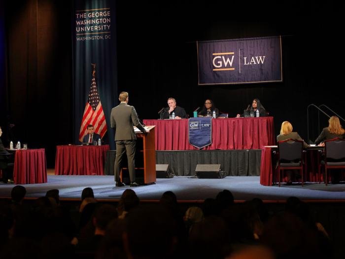 GW Law student William Schubert stands at a podium on a stage in front of a table of judges during the annual Van Vleck moot court competition.