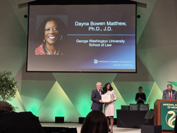 GW Law Dean Dayna Bowen Matthew accepts a diploma on stage during the 2025 NAM induction ceremony.