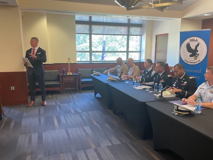 A group of military people sitting on a panel and a man standing up addressing the crowd
