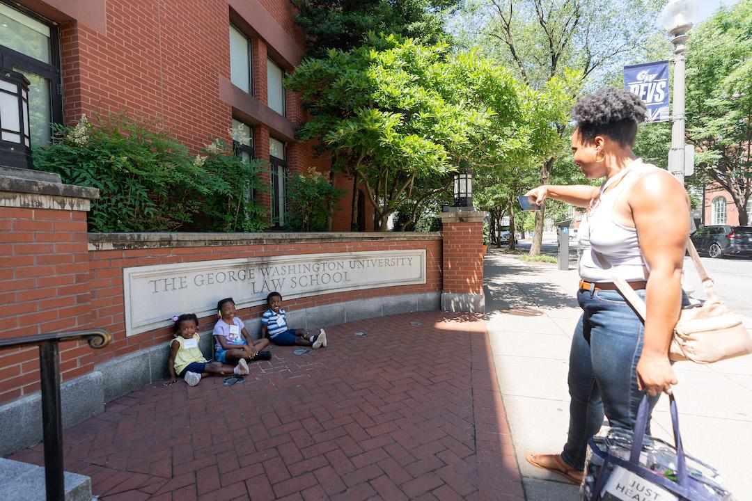 A woman taking a photo of three kids in front of the GW Law street sign