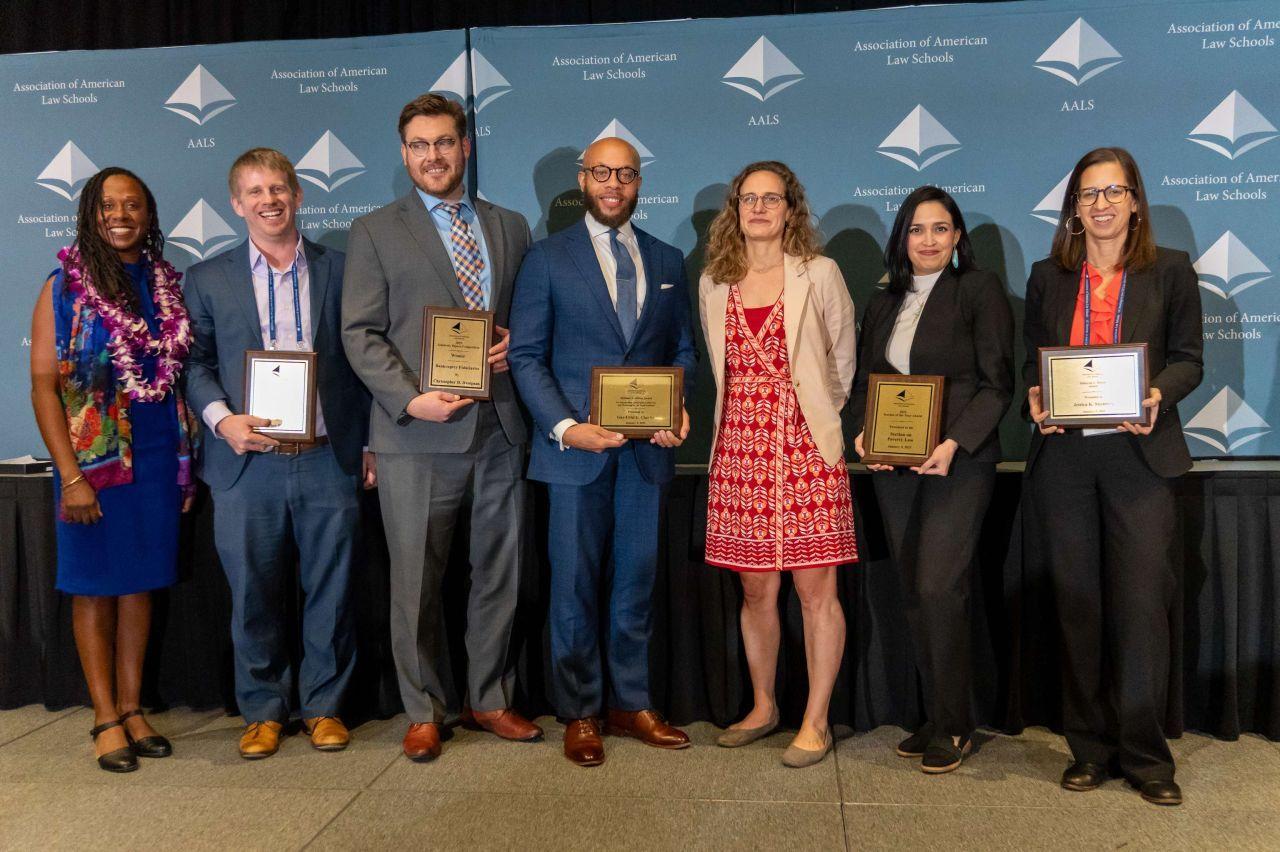 GW Law Professor Jessica Steinberg poses with other award winners at the 2025 AALS meeting. There are seven people in the photo lined up with award plaques in their hands in front of an AALS background.