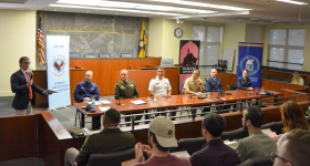 military personnel sitting at a table looking at the moderator speaking with a microphone to a crowd of students