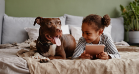 Front view portrait of cute African-American girl lying on bed with big pet dog and smiling, copy space